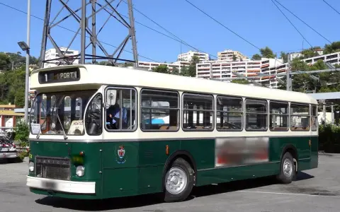 Un bus Saviem SC10, vert et crème, circule sous un ciel bleu. Il est stationné sur un parking, avec des immeubles en arrière-plan.