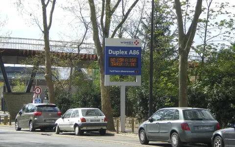 Panneau lumineux Vinci Autoroutes pour le Duplex A86, indiquant les tarifs. Des voitures sont garées sur le bord de la route.