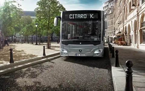 Un bus Mercedes-Benz Citaro K gris, dont le poids est impressionnant, roule dans une rue pavée bordée de bâtiments haussmanniens et d'arbres.