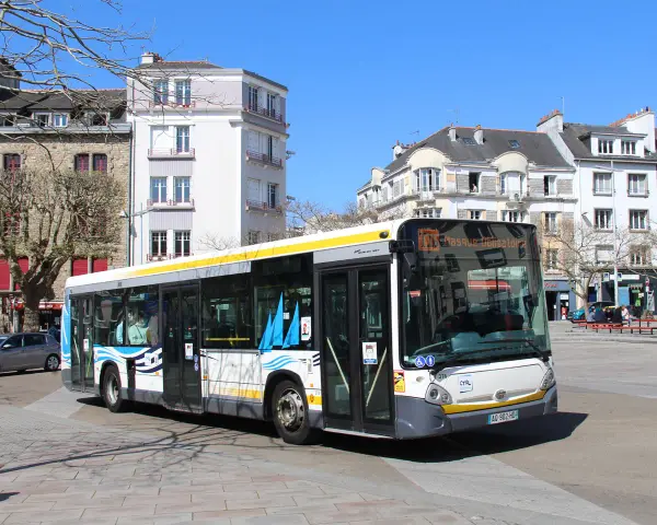 Un bus Heuliez GX 327 blanc et jaune circule dans une rue pavée, devant des immeubles anciens sous un ciel bleu.