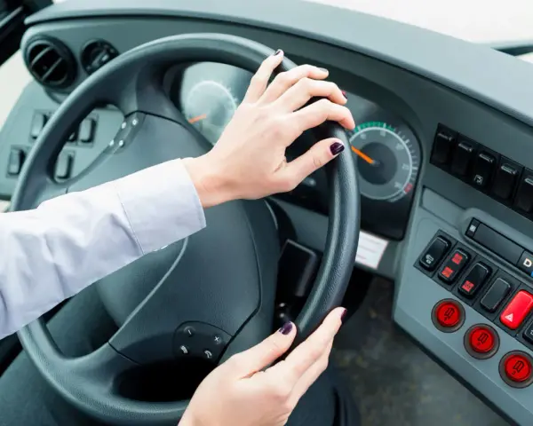 Vue de l'intérieur d'un bus, mains sur le volant. Le tableau de bord est visible, avec des boutons et des indicateurs. Le chauffeur est prêt à partir.