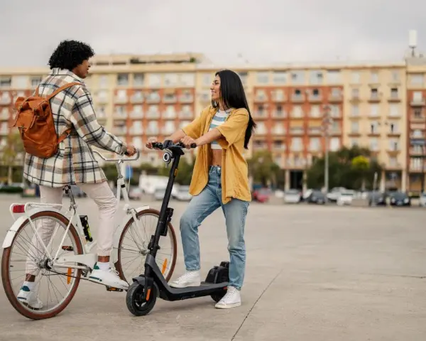 Deux jeunes femmes discutent, l'une sur un vélo blanc, l'autre sur une trottinette électrique. Elles profitent d'un moyen de transport doux en ville.