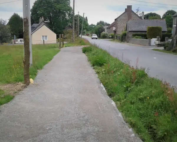 Un chemin de gravier longe une route de campagne bordée d'herbes folles. C'est un exemple de cheminement doux, invitant à la promenade paisible.