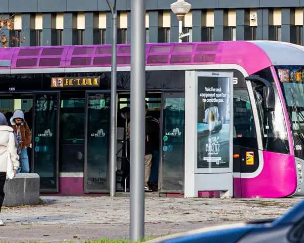 Un tramway rose et gris, un des transports en commun de Metz, s'arrête à un arrêt. Des passagers montent et descendent.