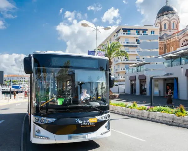 Un bus Esterel côte d'Azur circule dans une ville ensoleillée, près d'un port et d'une église.