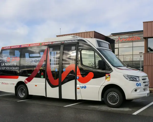 Un bus blanc avec des motifs rouges et noirs, portant l'inscription "Citéa La Navette" et des noms de localités, est garé devant un bâtiment "Valence Romans Mobilités".
