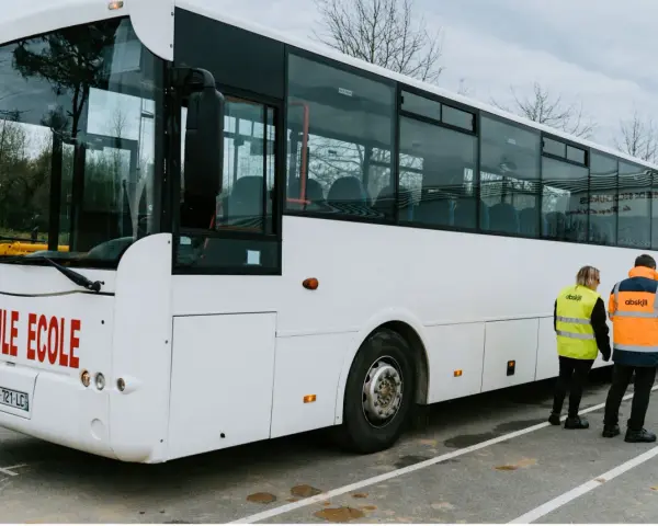 Un bus blanc "Véhicule École" est garé. Deux instructeurs, un homme et une femme, portent des gilets de haute visibilité.