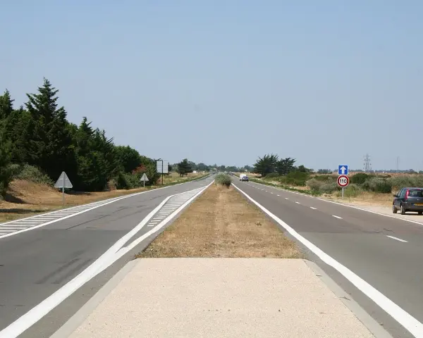 Une longue voie publique s'étend sous un ciel bleu clair. Des arbres bordent la route, et une voiture est garée sur le bas-côté.
