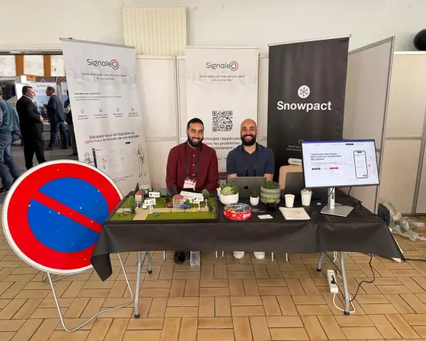 Deux hommes souriants présentent une maquette de ville et un panneau "Interdit de stationner" pour illustrer leur solution de **service voirie** connectée.