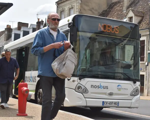 Un homme attend le bus à Nogent-le-Rotrou, sac à la main. Le panneau lumineux indique "NOBUS".