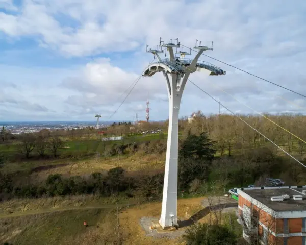 Un pylône blanc massif soutient un téléphérique urbain au-dessus d'une ville et d'une colline boisée.