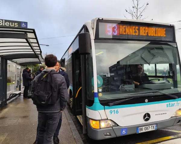 Un bus de transports urbains briochins s'arrête à un arrêt. Des passagers attendent, certains montent. Le panneau indique "53 Rennes République".