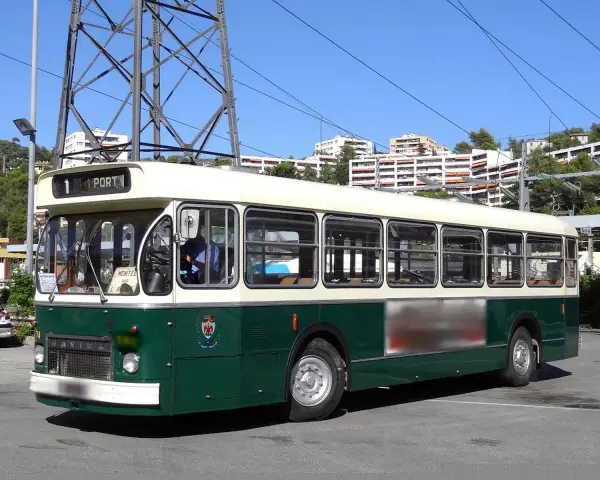 Un bus Saviem SC10, vert et crème, circule sous un ciel bleu. Il est stationné sur un parking, avec des immeubles en arrière-plan.