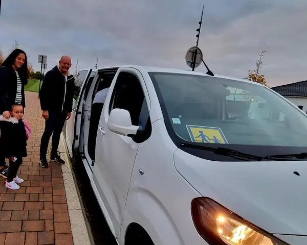 Famille attendant le transport scolaire communal. Le panneau jaune sur le pare-brise indique la présence d'enfants.