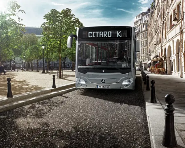 Un bus Mercedes-Benz Citaro K gris, dont le poids est impressionnant, roule dans une rue pavée bordée de bâtiments haussmanniens et d'arbres.
