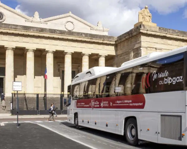 Un bus blanc "car régionaux Gironde" roule devant un bâtiment imposant avec des colonnes. Le bus arbore le slogan "Je roule au bipgaz".