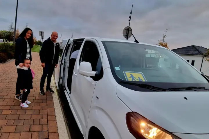 Famille attendant le transport scolaire communal. Le panneau jaune sur le pare-brise indique la présence d'enfants.