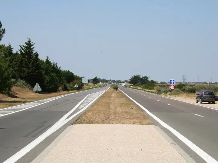 Une longue voie publique s'étend sous un ciel bleu clair. Des arbres bordent la route, et une voiture est garée sur le bas-côté.