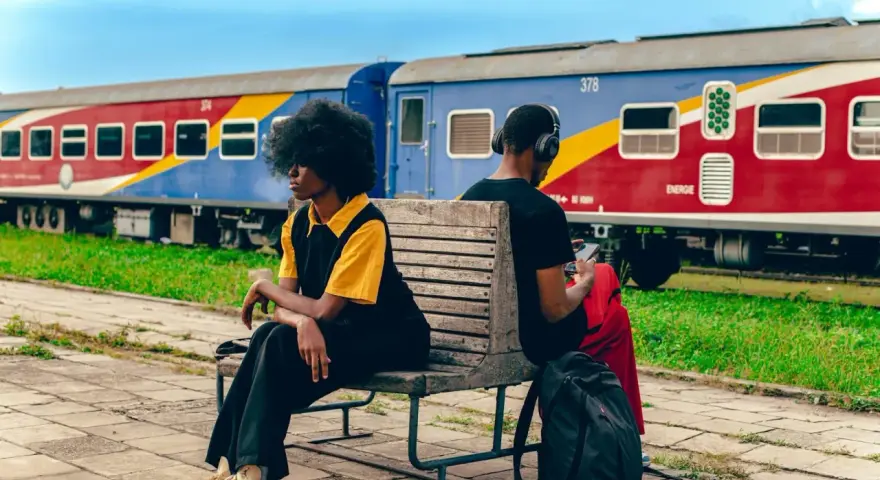 Deux personnes attendent sur un banc, un train coloré, un moyen de transport en Afrique, stationne derrière elles.