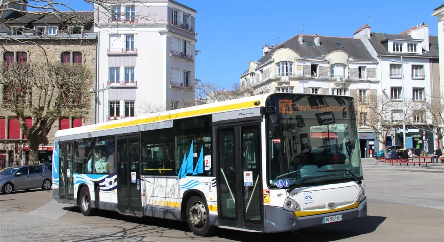 Un bus Heuliez GX 327 blanc et jaune circule dans une rue pavée, devant des immeubles anciens sous un ciel bleu.