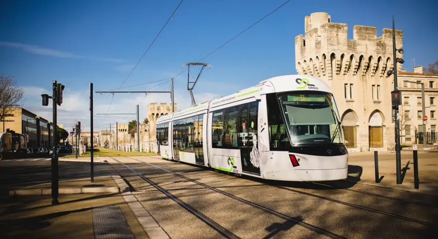 Le tramway moderne circule sur les rails à Avignon, passant devant des bâtiments historiques sous un ciel bleu éclatant.