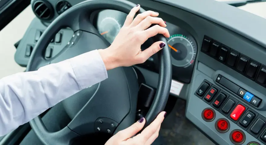 Vue de l'intérieur d'un bus, mains sur le volant. Le tableau de bord est visible, avec des boutons et des indicateurs. Le chauffeur est prêt à partir.