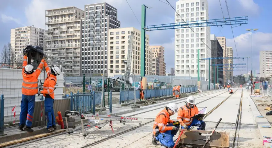 Des ouvriers travaillent sur de nouvelles infrastructures de tramway, avec des immeubles modernes en arrière-plan.