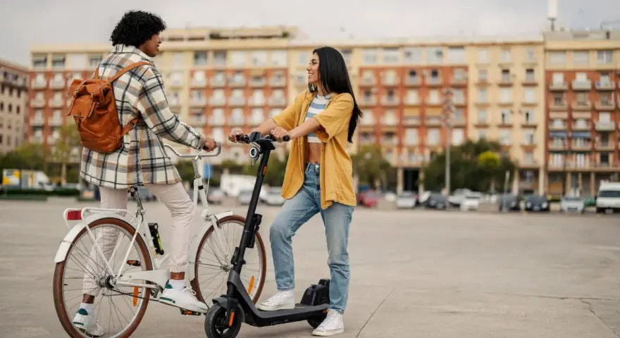 Deux jeunes femmes discutent, l'une sur un vélo blanc, l'autre sur une trottinette électrique. Elles profitent d'un moyen de transport doux en ville.