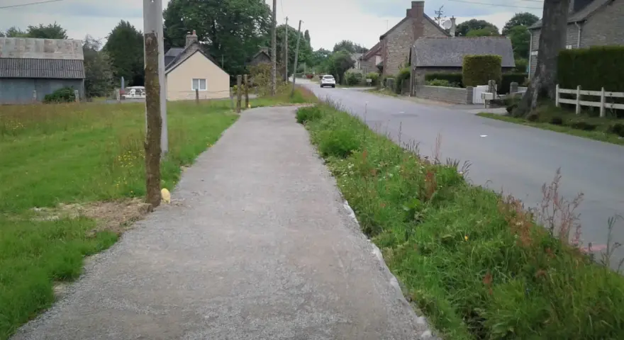 Un chemin de gravier longe une route de campagne bordée d'herbes folles. C'est un exemple de cheminement doux, invitant à la promenade paisible.