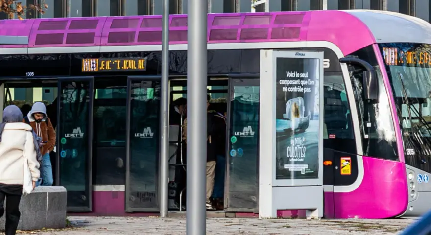 Un tramway rose et gris, un des transports en commun de Metz, s'arrête à un arrêt. Des passagers montent et descendent.