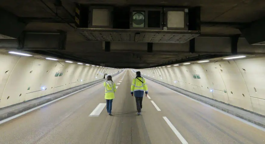 Deux personnes en gilets jaunes traversent un tunnel duplex de l'A86, la hauteur impressionnante du plafond est soulignée par les équipements techniques.