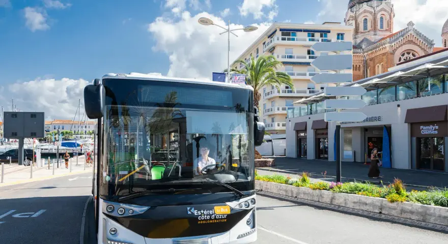 Un bus Esterel côte d'Azur circule dans une ville ensoleillée, près d'un port et d'une église.