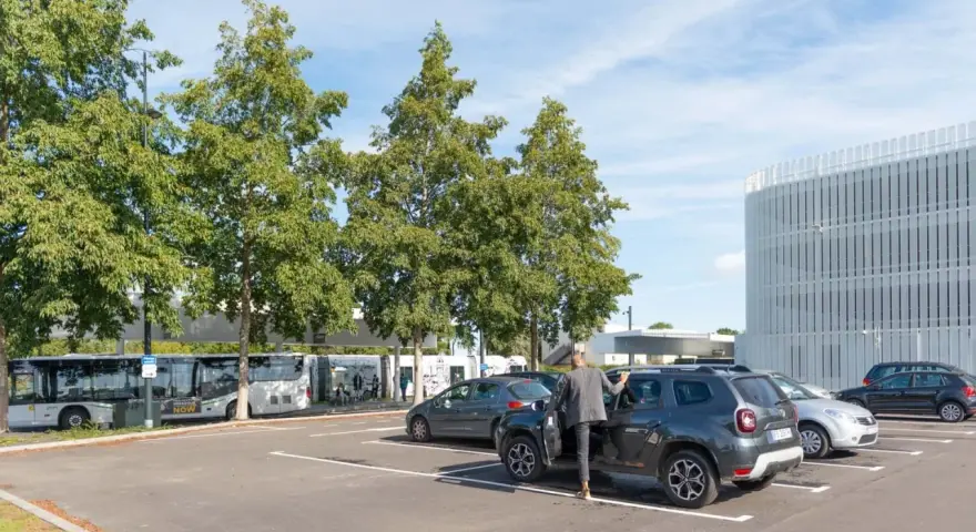 Un homme en costume monte dans un SUV gris dans un parking. Des bus électriques sont garés à proximité, près d'un bâtiment moderne. C'est une scène typique de naolib Nantes.