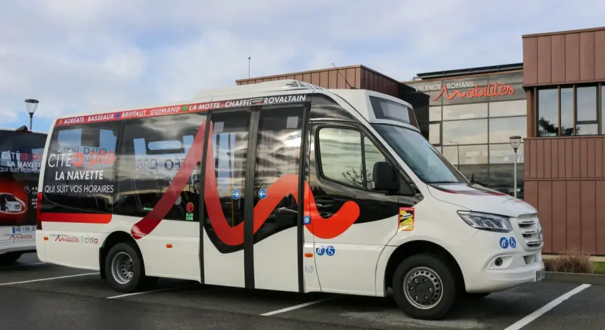 Un bus blanc avec des motifs rouges et noirs, portant l'inscription "Citéa La Navette" et des noms de localités, est garé devant un bâtiment "Valence Romans Mobilités".