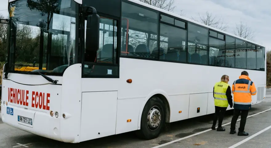 Un bus blanc "Véhicule École" est garé. Deux instructeurs, un homme et une femme, portent des gilets de haute visibilité.