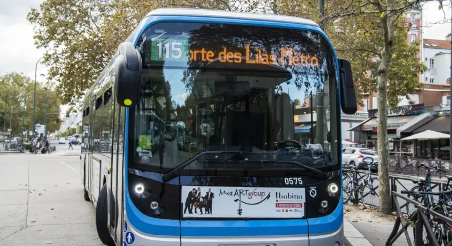 Bus Bolloré 115, direction Porte des Lilas Métro, stationné dans une rue bordée d'arbres et de commerces.