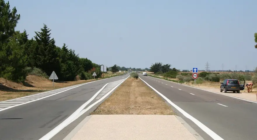 Une longue voie publique s'étend sous un ciel bleu clair. Des arbres bordent la route, et une voiture est garée sur le bas-côté.