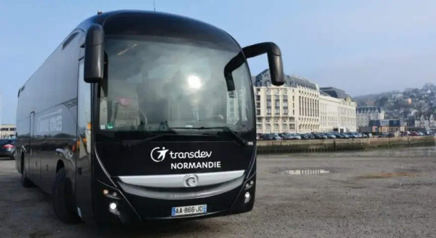 Autocar noir Transdev Normandie garé sur un quai, avec des bâtiments en arrière-plan.