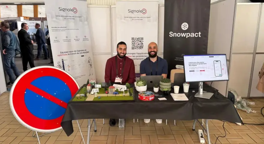 Deux hommes souriants présentent une maquette de ville et un panneau "Interdit de stationner" pour illustrer leur solution de **service voirie** connectée.