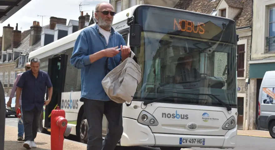 Un homme attend le bus à Nogent-le-Rotrou, sac à la main. Le panneau lumineux indique "NOBUS".