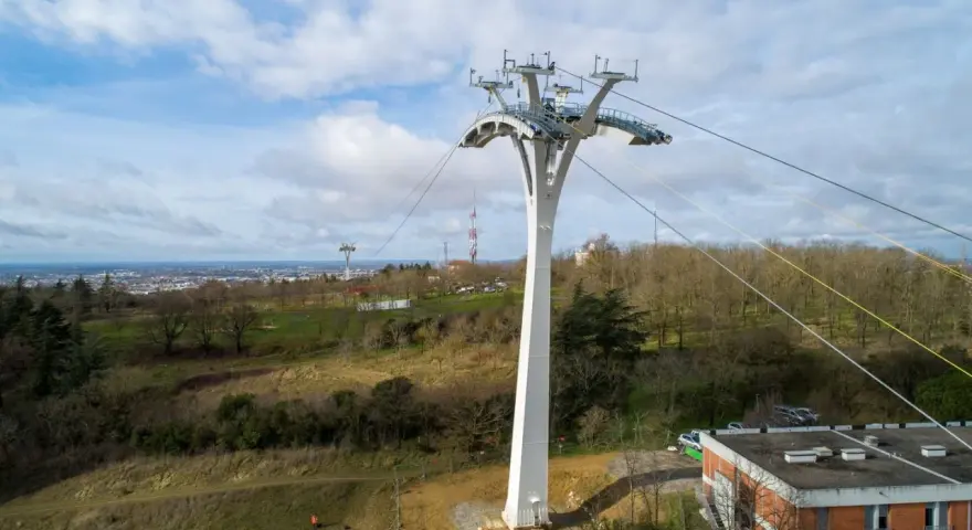 Un pylône blanc massif soutient un téléphérique urbain au-dessus d'une ville et d'une colline boisée.