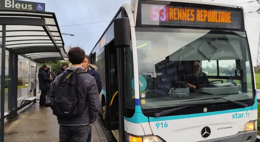 Un bus de transports urbains briochins s'arrête à un arrêt. Des passagers attendent, certains montent. Le panneau indique "53 Rennes République".