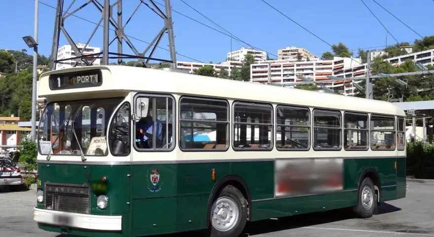 Un bus Saviem SC10, vert et crème, circule sous un ciel bleu. Il est stationné sur un parking, avec des immeubles en arrière-plan.