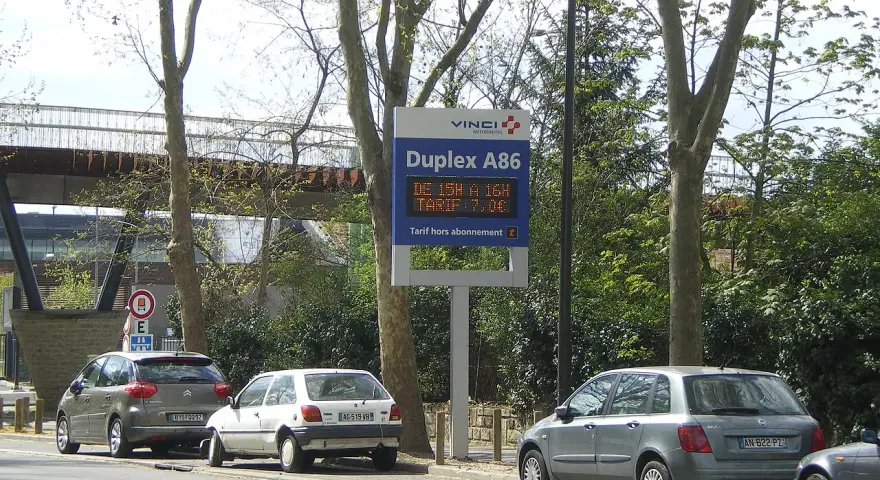 Panneau lumineux Vinci Autoroutes pour le Duplex A86, indiquant les tarifs. Des voitures sont garées sur le bord de la route.