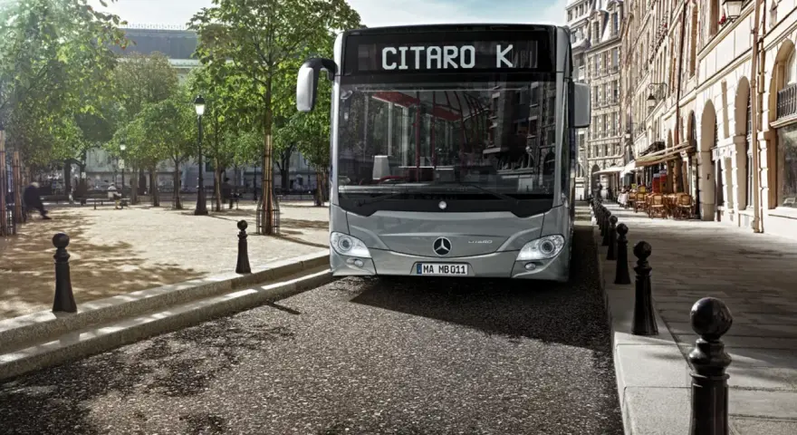 Un bus Mercedes-Benz Citaro K gris, dont le poids est impressionnant, roule dans une rue pavée bordée de bâtiments haussmanniens et d'arbres.