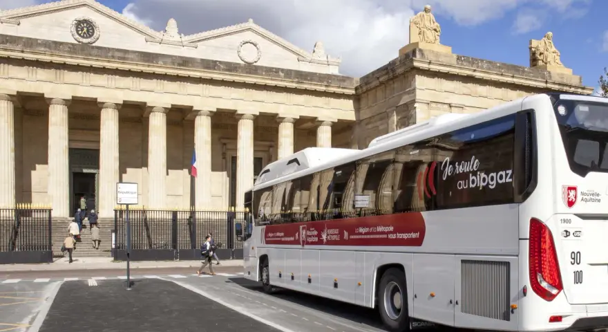 Un bus blanc "car régionaux Gironde" roule devant un bâtiment imposant avec des colonnes. Le bus arbore le slogan "Je roule au bipgaz".