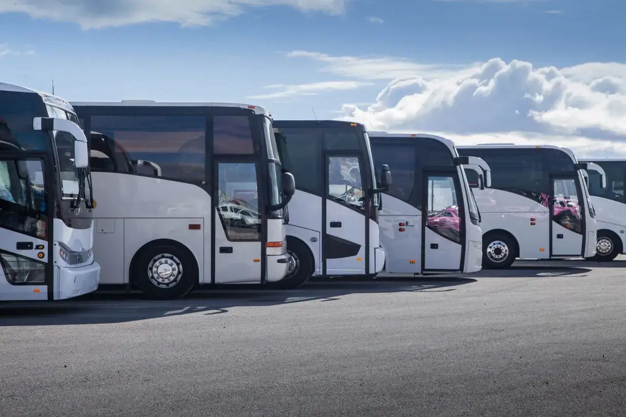 Une rangée de bus blancs attendent dans un parking sous un ciel bleu avec des nuages.