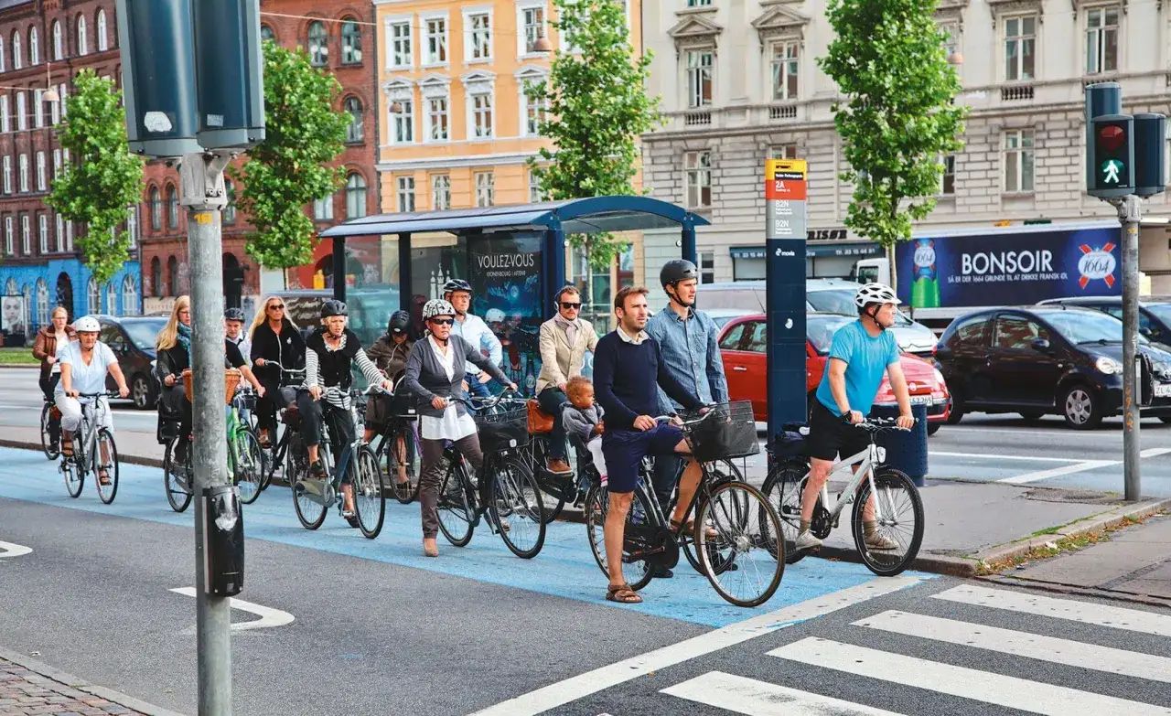 Une rue animée où plusieurs personnes utilisent le vélo comme moyen de transport doux. Des cyclistes de tous âges traversent la chaussée, profitant d'une journée ensoleillée.