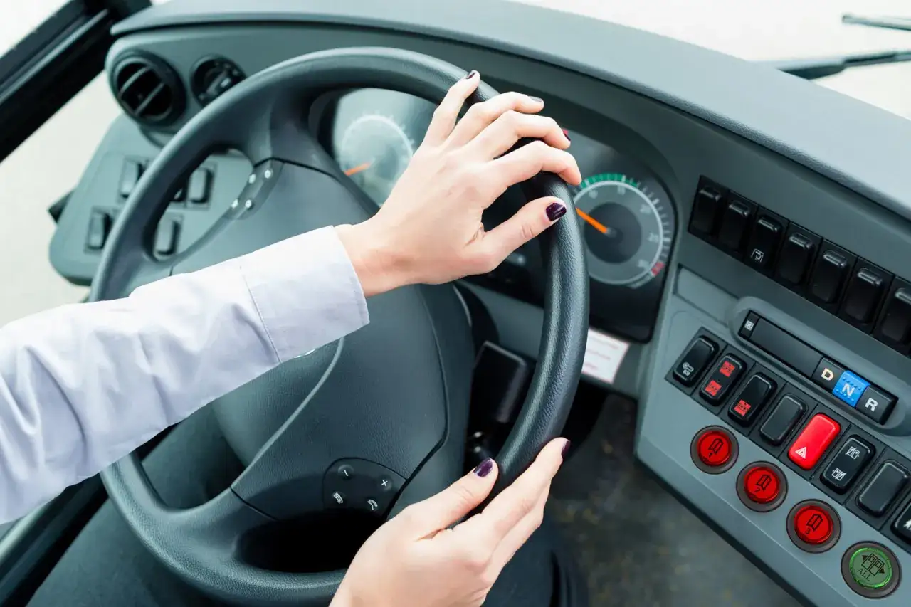 Vue de l'int&eacute;rieur d'un bus, mains sur le volant. Le tableau de bord est visible, avec des boutons et des indicateurs. Le chauffeur est pr&ecirc;t &agrave; partir.