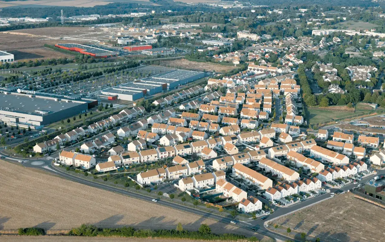 Vue aérienne d'une couronne périurbaine avec des maisons blanches aux toits orangés, des zones commerciales et des champs.